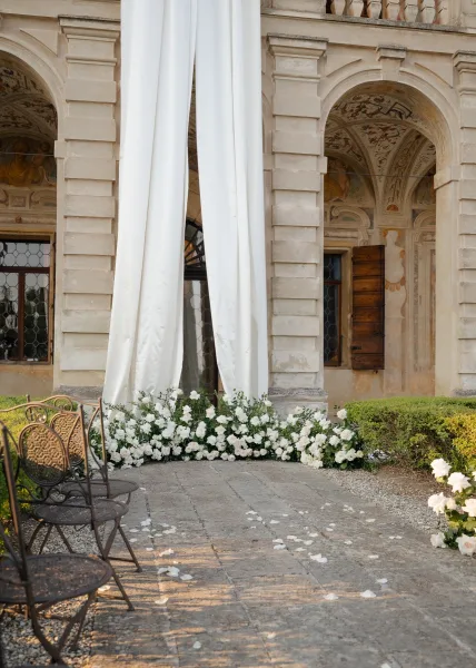 Ceremony setup with a statement drapery backdrop beneath the stone arches of a historic facade, along a stone walkway and garden hedges.