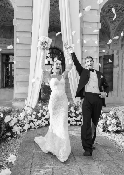 Ceremony exit amid a rose petal toss beneath a stone archway, bride in a lace bridal gown and groom walking the courtyard path together.
