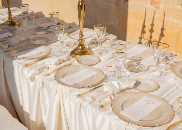 Reception tablescape in a gold and ivory table setting, highlighted by gold-rim glassware against a sunlit stucco wall with soft shadows.