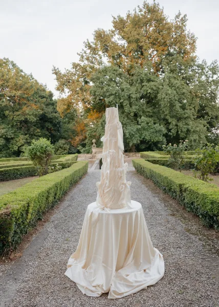 Tall wedding cake with fondant ruffles set against formal garden hedges and a gravel pathway, with trees rising beneath the sky.
