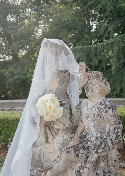 Bridal details arranged for a wedding detail photo, with a lace veil draped by a stone statue amid garden greenery and boxwood hedges.