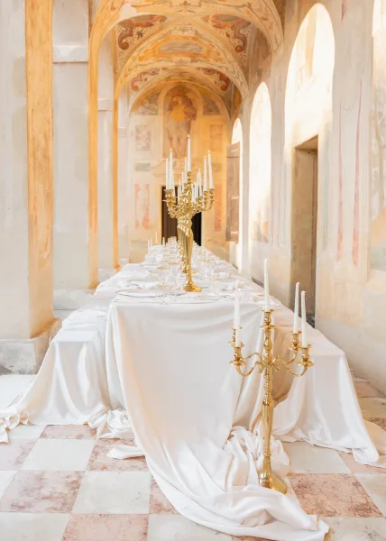 Reception tablescape set along a long banquet table with gold candelabras, beneath frescoed arches in an arched corridor.