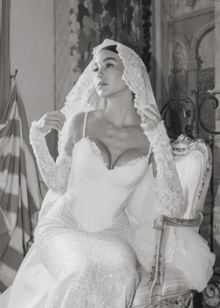 Bridal portrait of a bride adjusting her lace veil, posed against a painted wall mural with wrought iron detail and soft draped fabric.