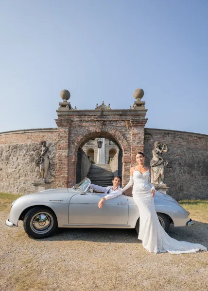 Wedding car portrait of a couple with a classic car, a vintage convertible, posed beneath a grand stone archway with statues and sunlit blue sky.