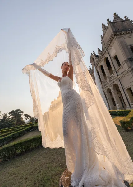 Bridal portrait of a bride with long veil in delicate gloves, poised on a manicured lawn before a grand mansion facade at sunset.