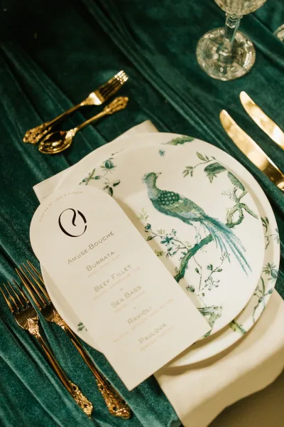 Reception tablescape on a green velvet tablecloth with gold flatware, patterned plate, printed menu card, white napkin, and wine glass