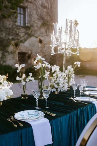 Reception tablescape with emerald velvet tablecloth, white florals and crystal candelabra, peacock feathers on a sunset-lit terrace near stone walls
