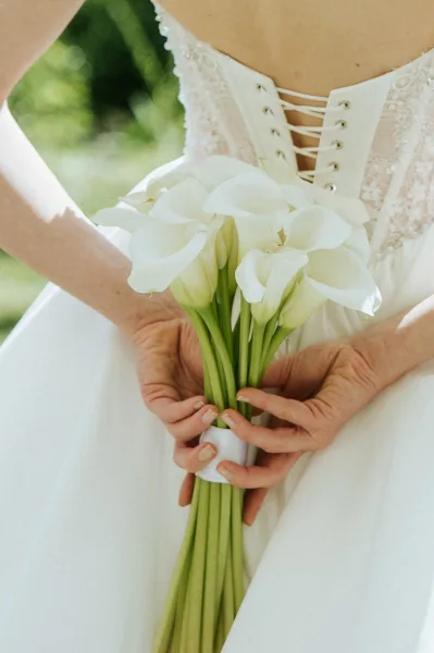 Bridal bouquet of white calla lilies tied with a ribbon wrap, held against a lace corset wedding dress with soft greenery bokeh behind