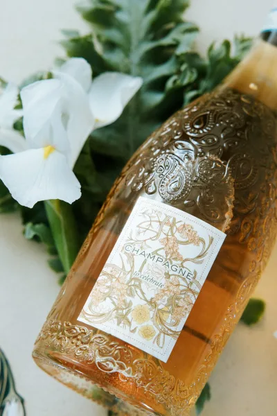 Champagne bottle close up with embossed glass and floral label, styled with a white flower, greenery, and cork on a light tabletop surface