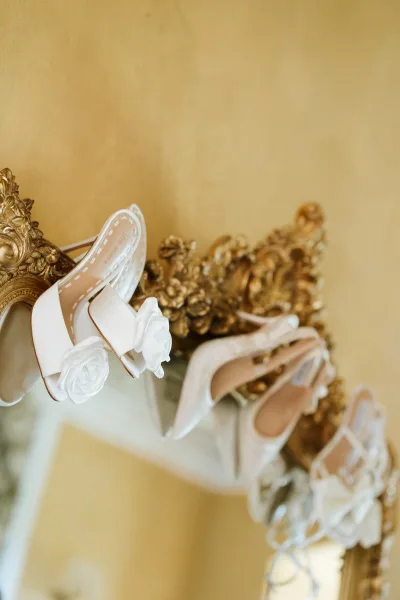 Bridal shoes, white wedding heels with rosette flower appliqués resting on an ornate gold mirror against a gold wall reflection
