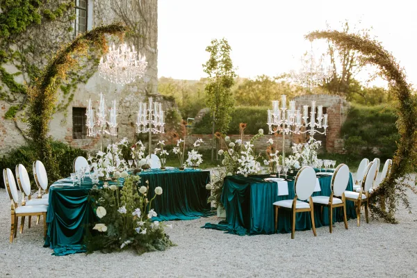 Reception tablescape with emerald velvet tablecloth, white floral centerpieces and crystal chandeliers in a gravel courtyard by stone walls