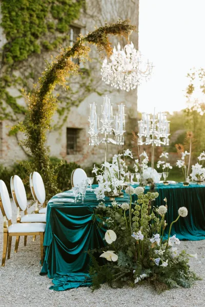 Reception tablescape with an emerald velvet tablecloth, crystal chandelier and glass candelabras, set in a stone courtyard garden at sunset