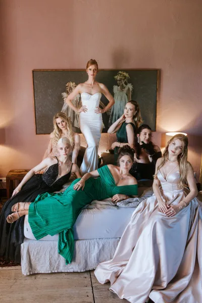 Bridesmaid group portrait of the bride with bridesmaids posing on a bed in a pink-walled bedroom, in satin dresses with statement earrings