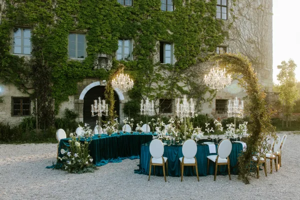 Reception tablescape with teal wedding tablecloth, white oval chairs, chandeliers and tall candelabras in an ivy-covered stone courtyard