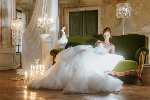 Bridal portrait of a bride reclining on a green velvet sofa in a strapless tulle gown, with chandelier glow against aged plaster walls
