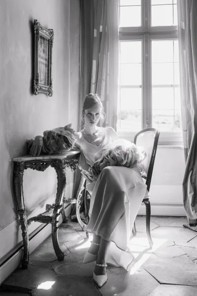 Bridal portrait in natural light, bride holding a bouquet while seated by a large window, wearing jewelry and pointed toe heels indoors.