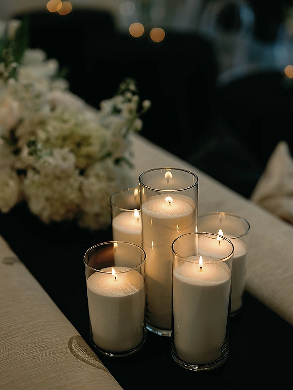 Wedding candle centerpiece with cylinder vase candles and white pillar candles, surrounded by florals on a runner with bokeh reception lights
