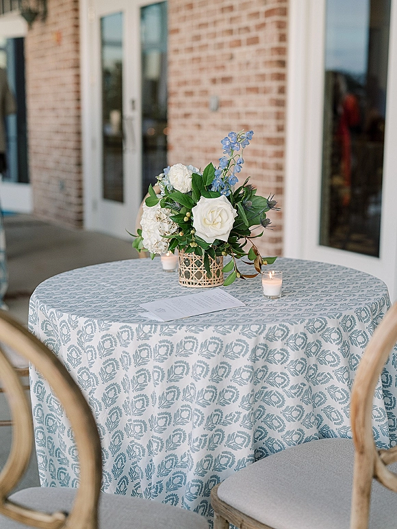 Reception tablescape with wedding cocktail table decor featuring white roses and blue delphinium in a woven vase, votive candles, menu card on a patio