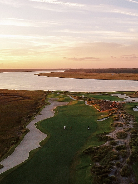 Landscape scenery with sunset river view over marshland at golden hour, showing golf course greens, sand bunkers, and an excavator near carts
