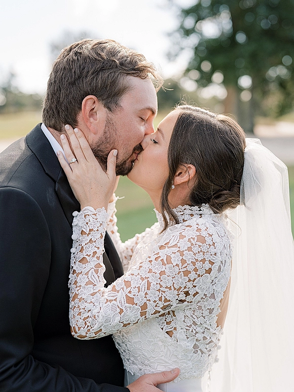 Wedding kiss portrait of bride and groom kissing as she holds his face, lace sleeve and engagement ring visible amid outdoor greenery