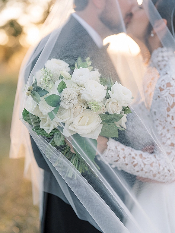 Wedding kiss portrait of bride and groom kissing under a veil, bride holding a white rose bouquet in golden hour greenery background