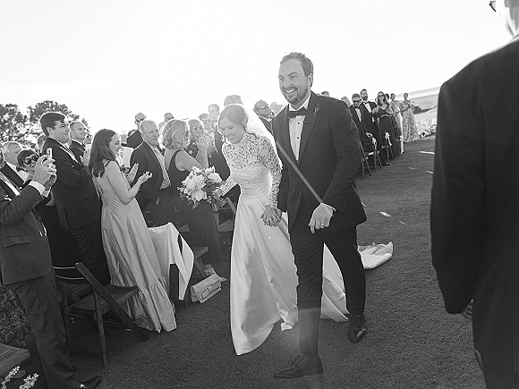Wedding recessional as bride and groom walk the aisle holding hands, bride with bouquet and veil, guests cheering on an outdoor lawn