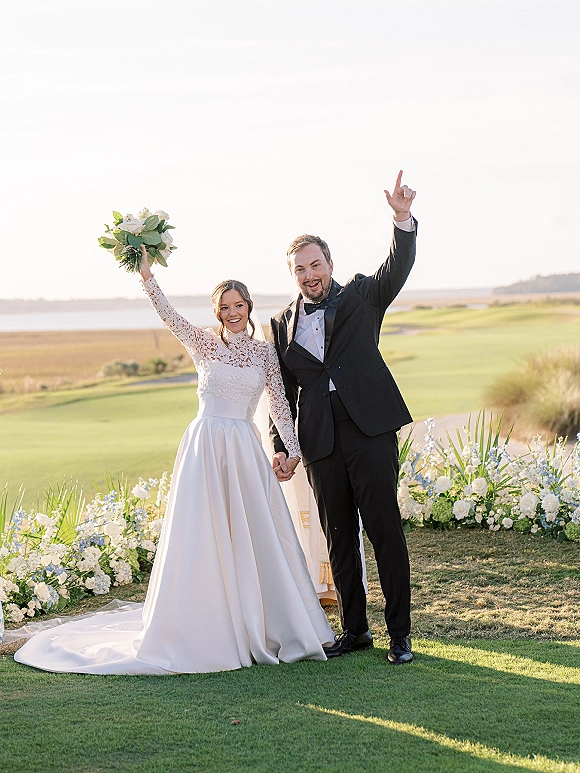 Ceremony recessional as newlyweds walk hand in hand down a floral-lined lawn aisle by the water, bride lifting bouquet, groom cheering in tuxedo