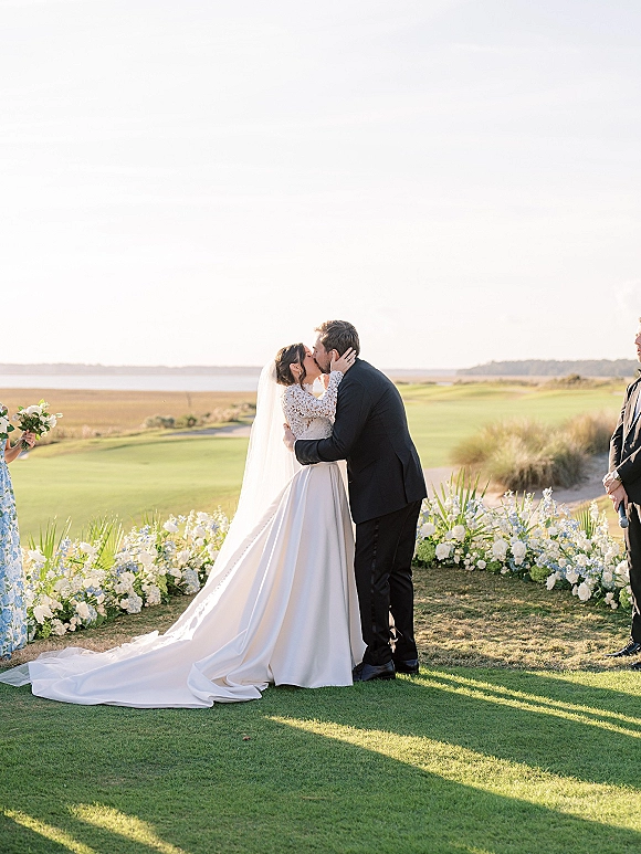 Wedding kiss at an outdoor wedding ceremony as the bride in a long veil holds the groom’s face on a lawn with ocean and dunes behind