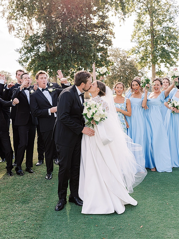 Wedding kiss portrait of the bride and groom kiss as their wedding party cheers, bride holding bouquet and veil on a sunny lawn with trees