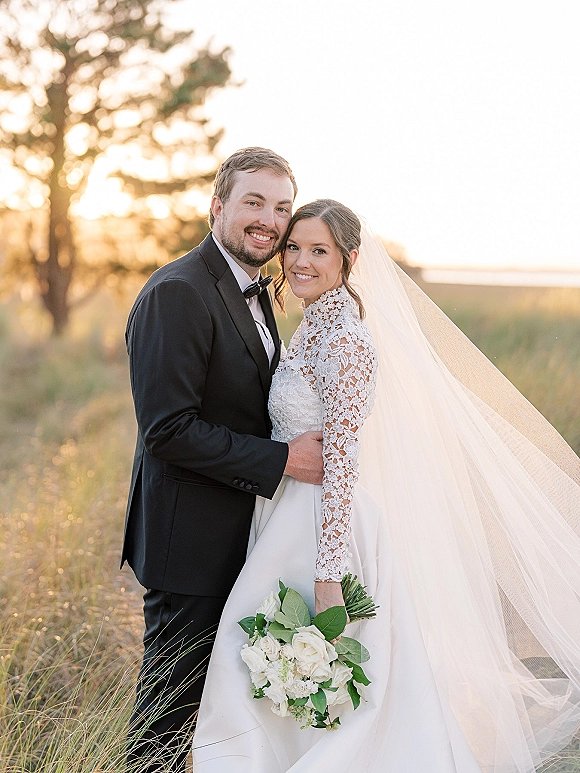 Couple portrait of bride and groom portrait embracing in a sunset field, her lace long-sleeve gown and veil flowing, holding white roses bouquet