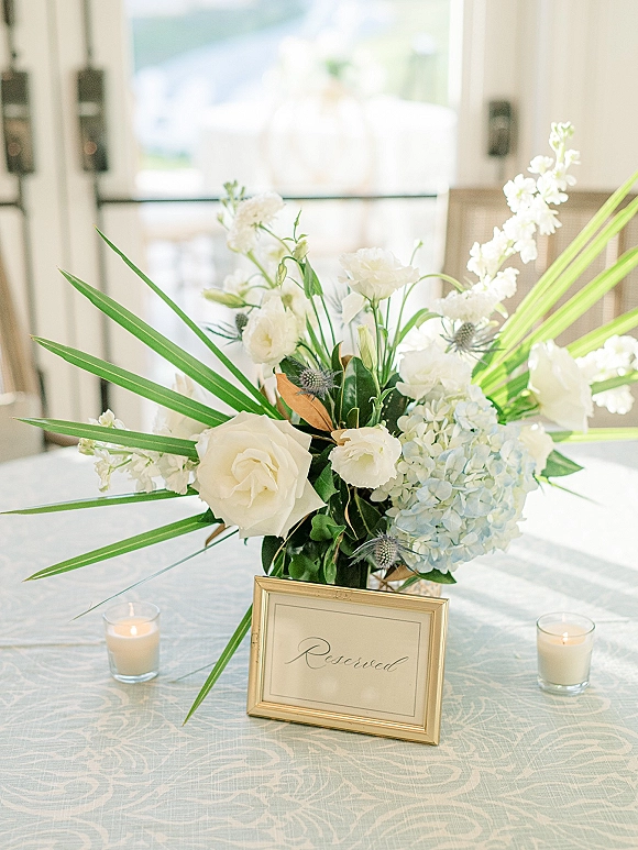 Wedding centerpiece with white and blue hydrangeas and roses in a tall arrangement beside a framed reserved sign and candles near a bright window