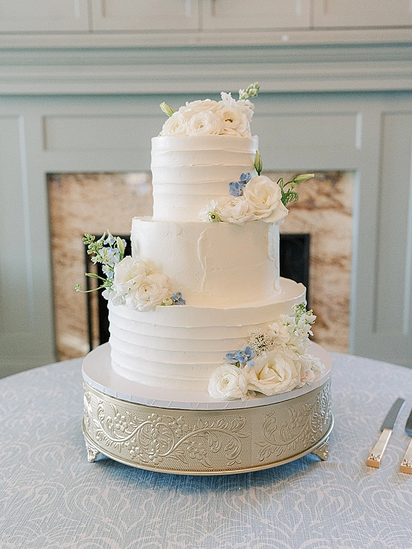 Wedding cake with smooth white buttercream frosting, three tiers topped with white roses and blue flowers on a silver stand by a stone fireplace mantel