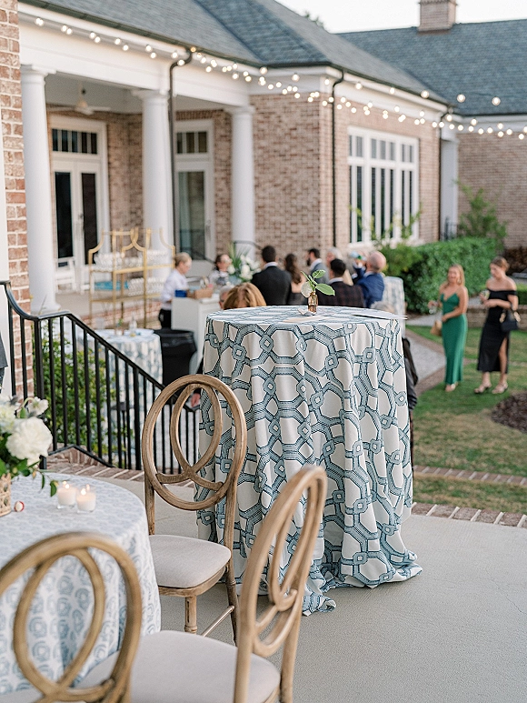 Cocktail hour decor with a wedding cocktail table in blue patterned linen, bud vase and votive candles under string lights on patio by brick building