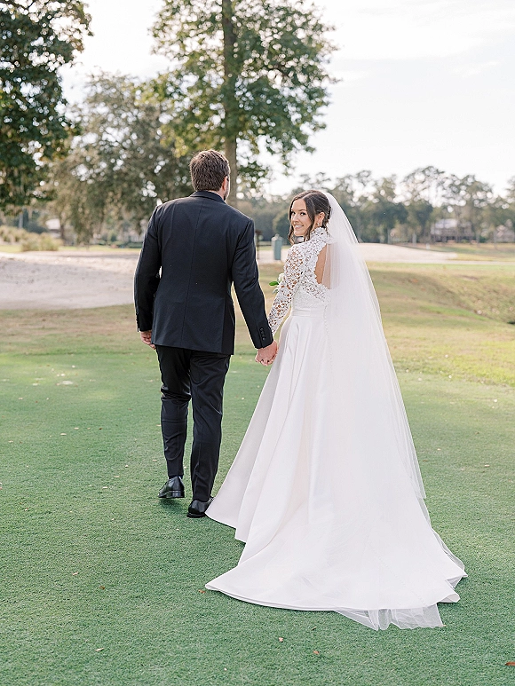 Couple portrait of bride and groom walking away holding hands, bride looking back in cathedral veil on a golf course lawn