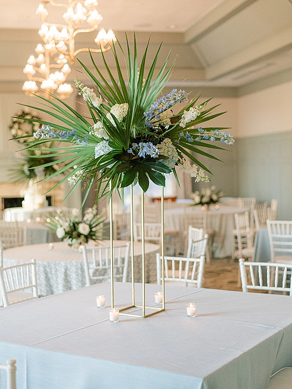 Wedding centerpiece with a tall wedding centerpiece of palm fronds and blue-white flowers on a gold geometric stand, with candles under a chandelier