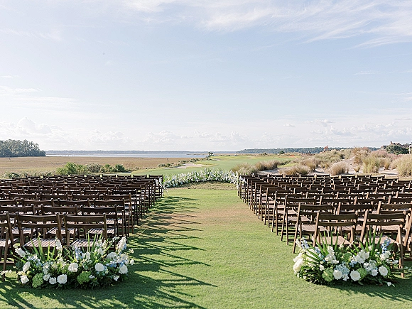 Ceremony setup with wood folding chairs lining a floral aisle, white and blue blooms on a lawn facing dunes and water horizon