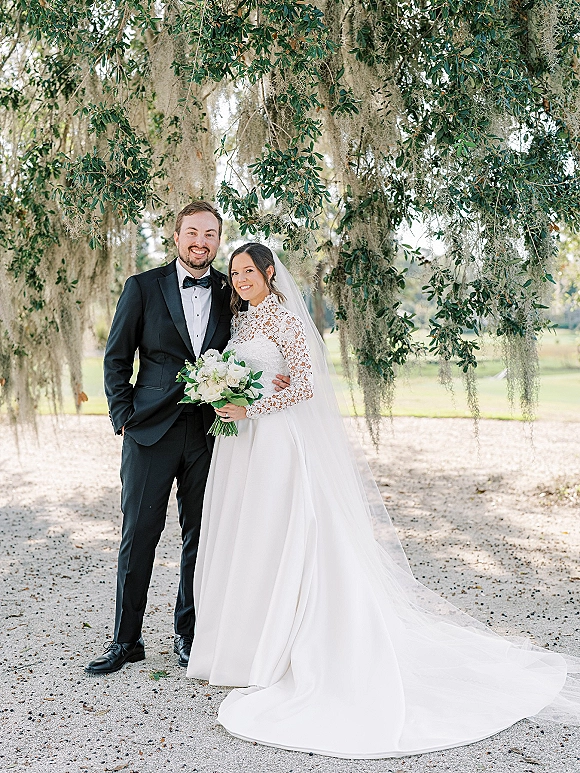 Couple portrait of bride in lace long-sleeve gown and veil holding white rose bouquet with greenery, groom in tux under oak with Spanish moss