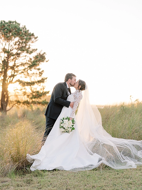 Wedding kiss portrait of bride and groom kiss, her cathedral veil and white rose bouquet flowing as they dip in a sunset field