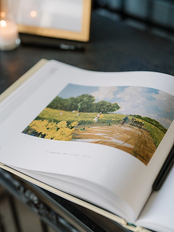 Wedding guest book open on a dark tabletop with printed landscape artwork, pen and candle in glass holder beside the pages