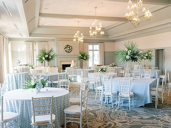 Reception tablescape in a wedding reception room with light blue linens, white chiavari chairs, floral centerpieces, and chandeliers above