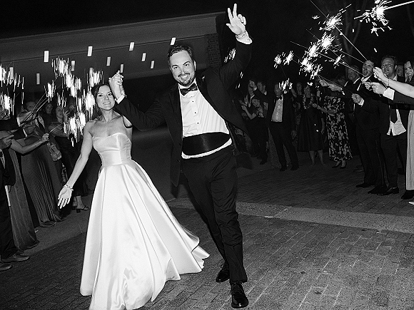 Sparkler exit as bride in a strapless satin dress and groom in tuxedo run through guests’ sparklers under string lights on a brick patio at night