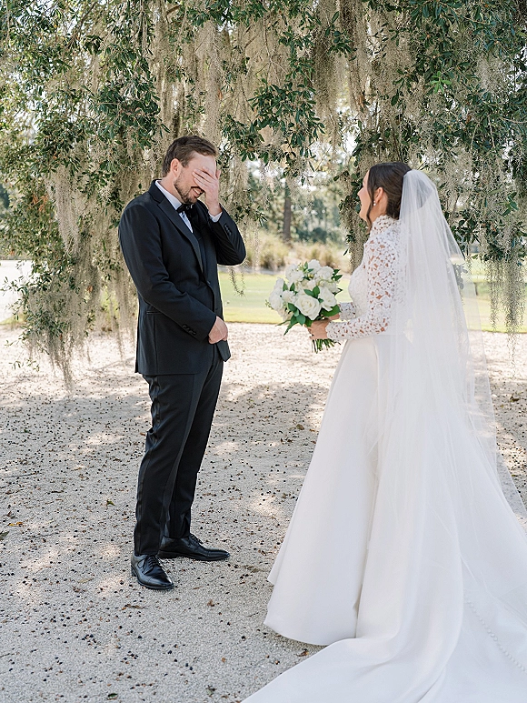 Wedding first look as the groom reaction first look unfolds under an oak tree with Spanish moss, bride in lace sleeves holding white roses