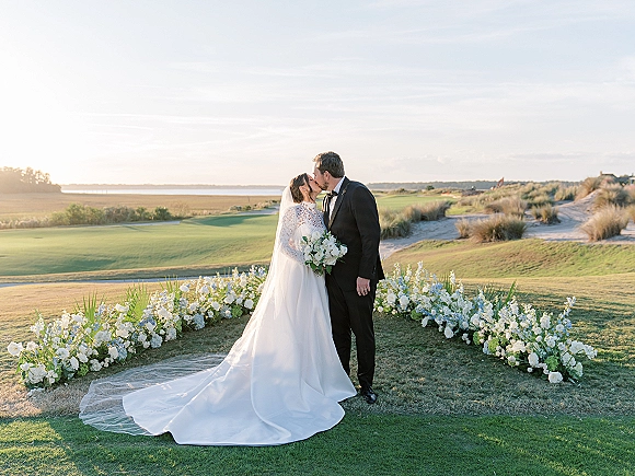 Wedding kiss portrait of bride and groom kissing, her long veil flowing as she holds a white rose bouquet on a coastal golf course lawn.