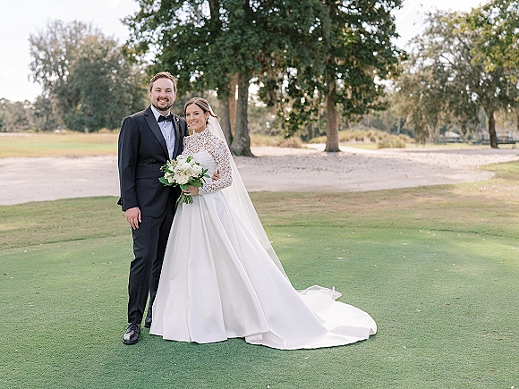 Couple portrait of bride in long sleeve lace gown with cathedral veil holding white rose bouquet beside groom in tux on golf course lawn