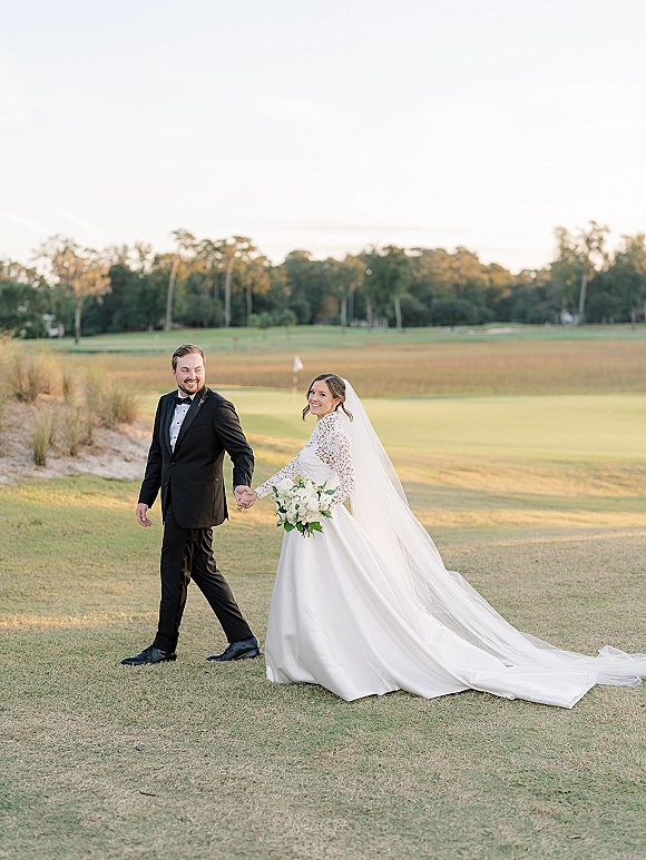 Couple portrait of bride and groom walking hand in hand on a golf course lawn, her cathedral veil and bouquet trailing behind him in a tuxedo