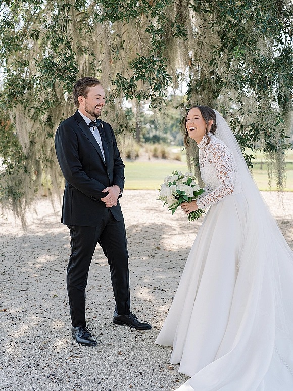 First look moment outdoors as bride in long sleeve lace dress and veil laughs at groom in black tux under oak tree with hanging moss