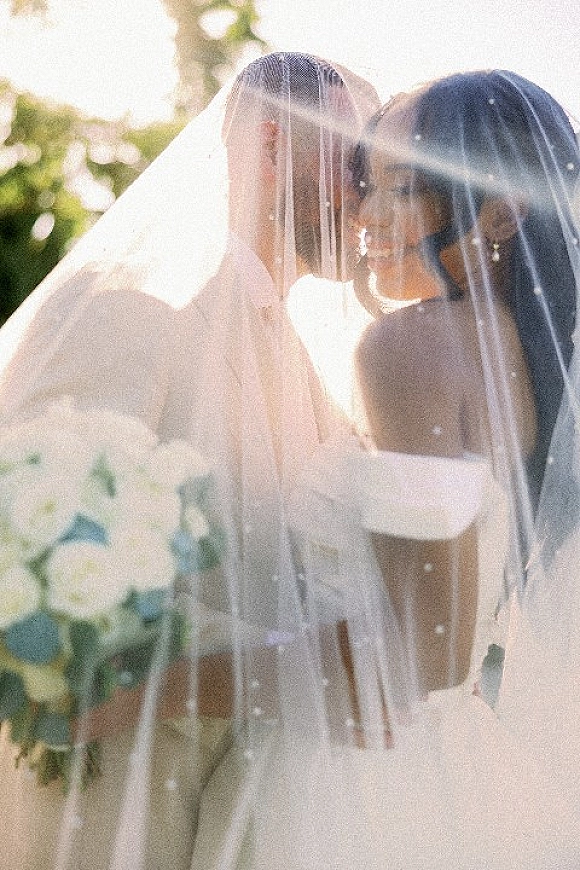 Wedding kiss portrait of couple kissing under a veil, bride holding a white bouquet as sunlight filters through outdoor greenery