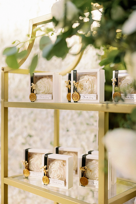 Wedding favors in clear box wedding favors packaging with preserved roses, black ribbon and wax seal tags on a gold shelf outdoors in soft light