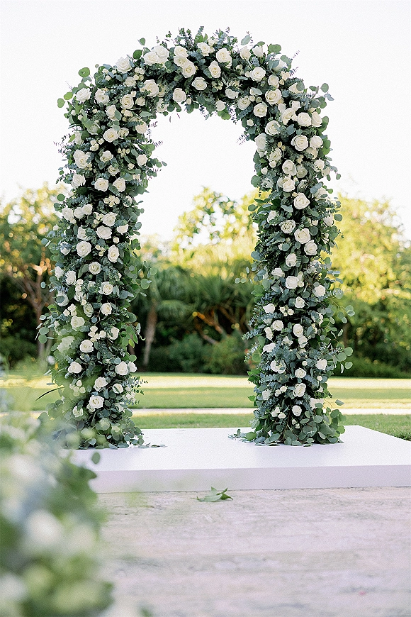 Wedding ceremony arch with white roses and eucalyptus greenery on a white platform stage, set on a garden lawn with trees behind