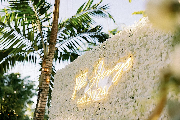 Wedding flower wall with neon wedding sign and string lights, a white floral backdrop set outdoors with palm trees and sky behind